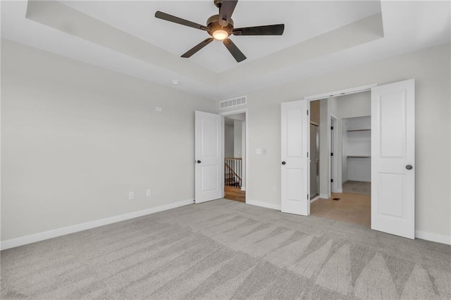 unfurnished bedroom with a raised ceiling, a walk in closet, light carpet, and ceiling fan