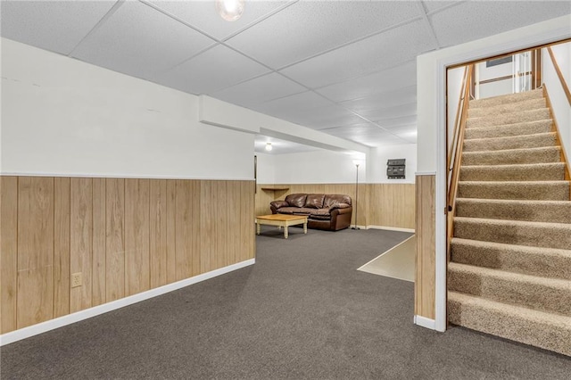 basement with a paneled ceiling, wooden walls, and dark colored carpet