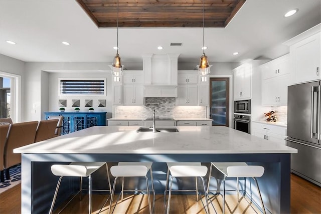 kitchen featuring decorative light fixtures, custom range hood, stainless steel appliances, and an island with sink