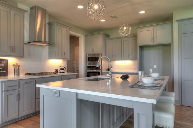 kitchen featuring pendant lighting, wall chimney exhaust hood, stainless steel appliances, and a kitchen island with sink