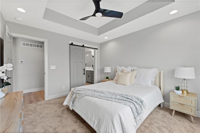 carpeted bedroom with a barn door, connected bathroom, ceiling fan, and a tray ceiling