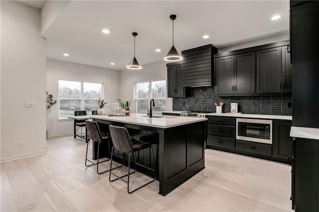 kitchen with built in microwave, wall chimney range hood, sink, decorative light fixtures, and an island with sink