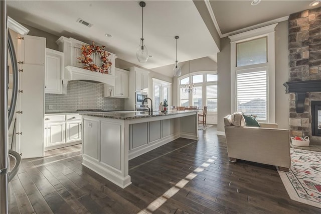 kitchen with a center island with sink, a stone fireplace, hanging light fixtures, dark stone countertops, and white cabinetry