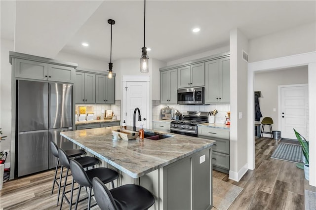 kitchen featuring hardwood / wood-style floors, an island with sink, appliances with stainless steel finishes, decorative light fixtures, and light stone counters