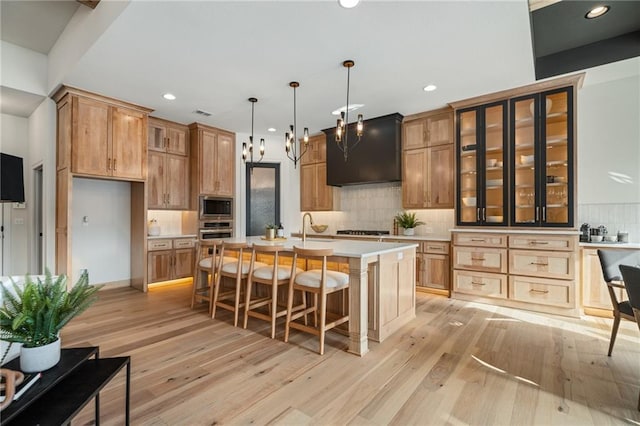 kitchen featuring light hardwood / wood-style flooring, hanging light fixtures, an island with sink, stainless steel appliances, and wall chimney exhaust hood