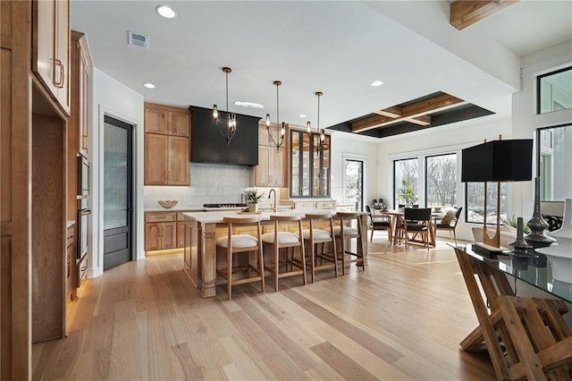 kitchen with decorative light fixtures, beam ceiling, decorative backsplash, light wood-type flooring, and an island with sink