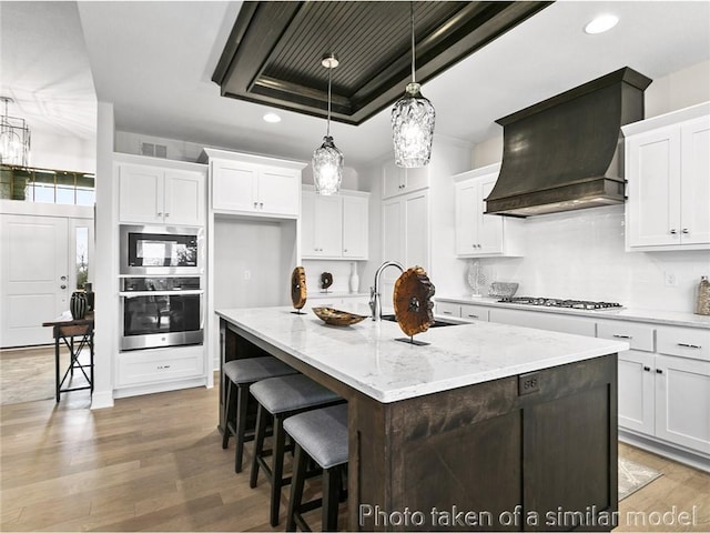 kitchen featuring premium range hood, a kitchen island with sink, hanging light fixtures, white cabinetry, and appliances with stainless steel finishes