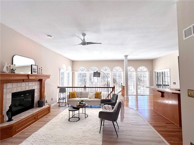 living room with visible vents, light wood-style flooring, a ceiling fan, a tile fireplace, and ornate columns