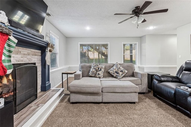 carpeted living room with a textured ceiling, a stone fireplace, and ceiling fan