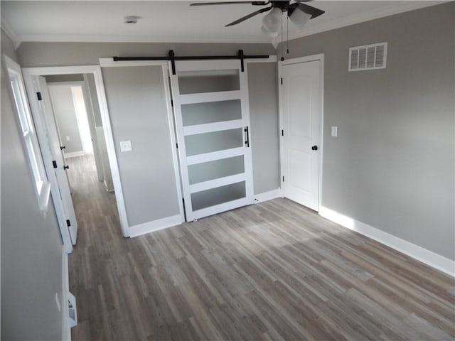 unfurnished bedroom with ceiling fan, a barn door, crown molding, wood-type flooring, and a closet