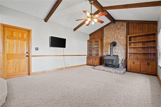 unfurnished living room with a ceiling fan, a wood stove, vaulted ceiling with beams, carpet, and a textured ceiling