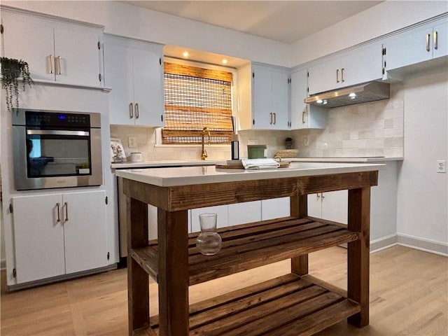 kitchen featuring light wood-type flooring, light countertops, under cabinet range hood, stainless steel oven, and tasteful backsplash