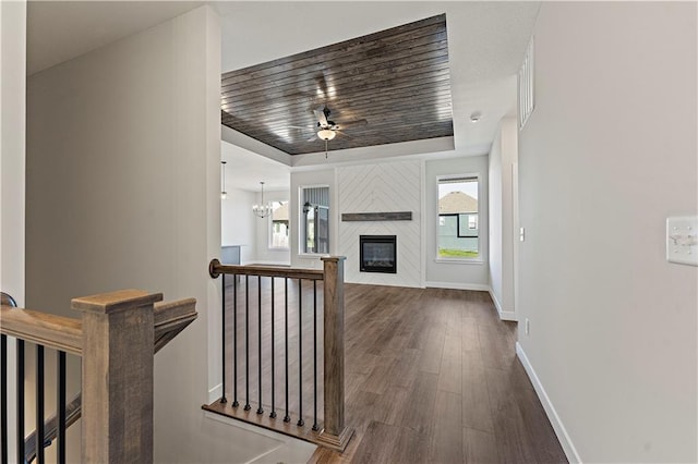 unfurnished living room featuring ceiling fan with notable chandelier, dark wood-style flooring, baseboards, a raised ceiling, and a glass covered fireplace