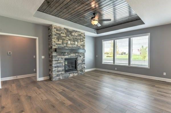 unfurnished living room with wood finished floors, baseboards, ceiling fan, a stone fireplace, and a raised ceiling