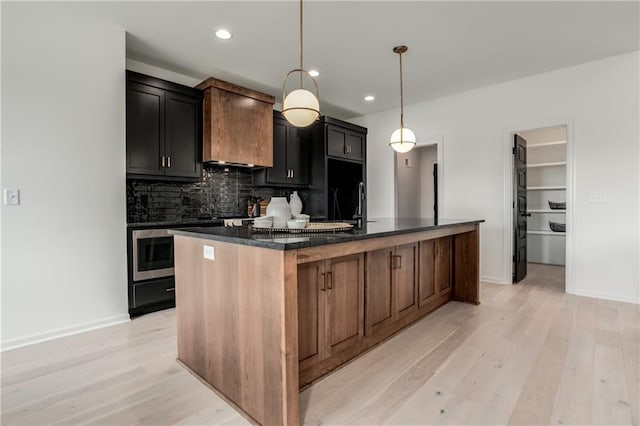 kitchen with light wood-type flooring, backsplash, dark stone counters, a kitchen island with sink, and decorative light fixtures