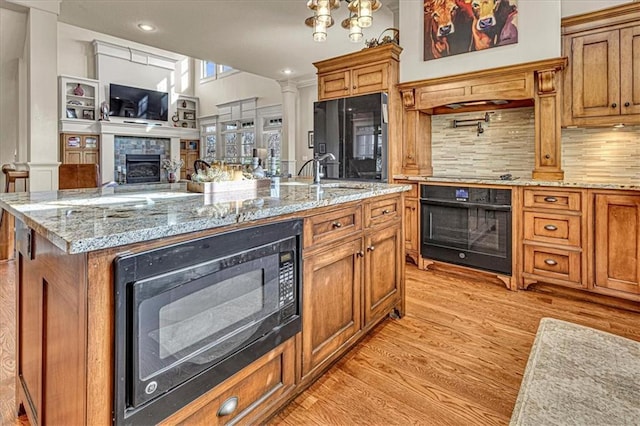 kitchen featuring a stone fireplace, an island with sink, decorative backsplash, black appliances, and light wood-type flooring