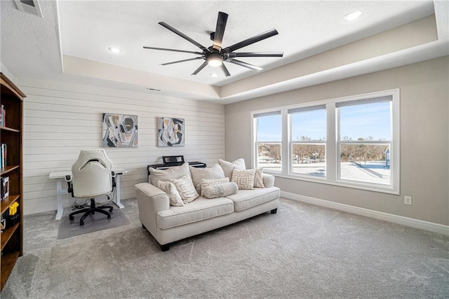 carpeted living room featuring ceiling fan, wooden walls, and a tray ceiling