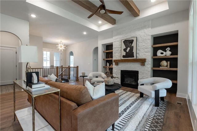 living room with beamed ceiling, light wood-type flooring, built in features, ceiling fan with notable chandelier, and a stone fireplace