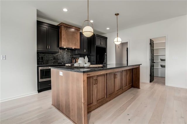 kitchen with light wood-type flooring, backsplash, dark stone counters, hanging light fixtures, and an island with sink