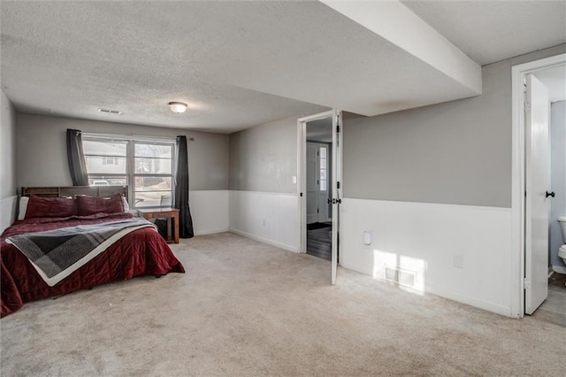 bedroom featuring light carpet, a textured ceiling, visible vents, and a wainscoted wall