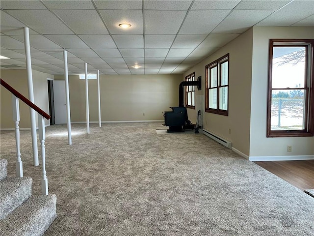 basement featuring a paneled ceiling, carpet, a wood stove, and a baseboard heating unit