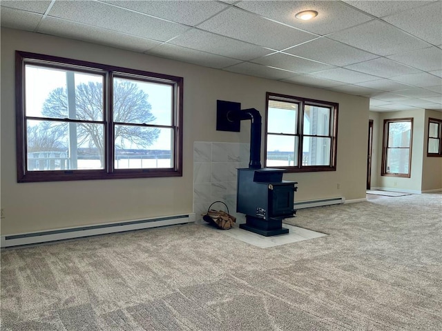 unfurnished living room featuring a drop ceiling, a baseboard radiator, a wood stove, and carpet