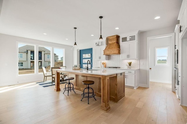 kitchen with white cabinetry, premium range hood, pendant lighting, light hardwood / wood-style floors, and a center island with sink