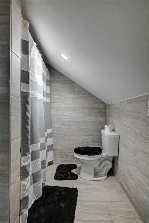 bathroom featuring toilet, a shower with shower curtain, vaulted ceiling, and tile walls