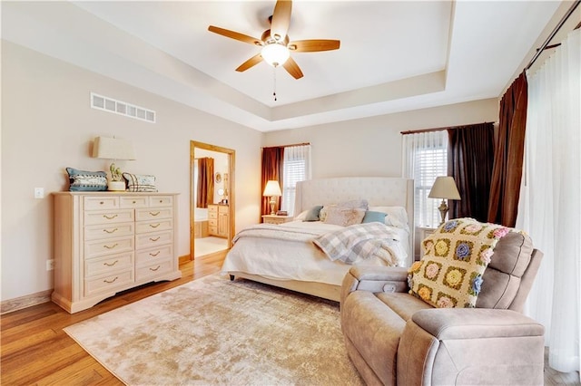 bedroom featuring a raised ceiling, ensuite bath, ceiling fan, and light wood-type flooring