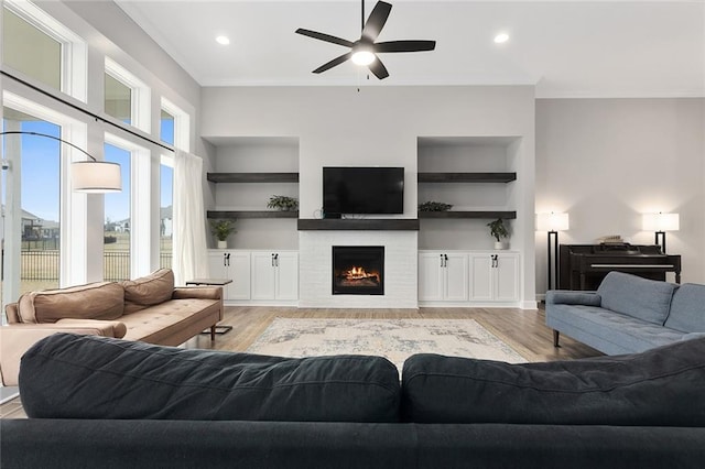 living area featuring crown molding, a lit fireplace, light wood finished floors, a ceiling fan, and built in shelves