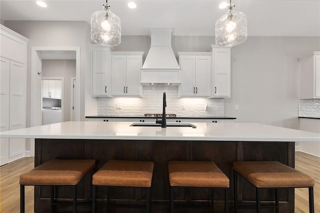 kitchen with custom exhaust hood, light wood finished floors, a center island with sink, and tasteful backsplash