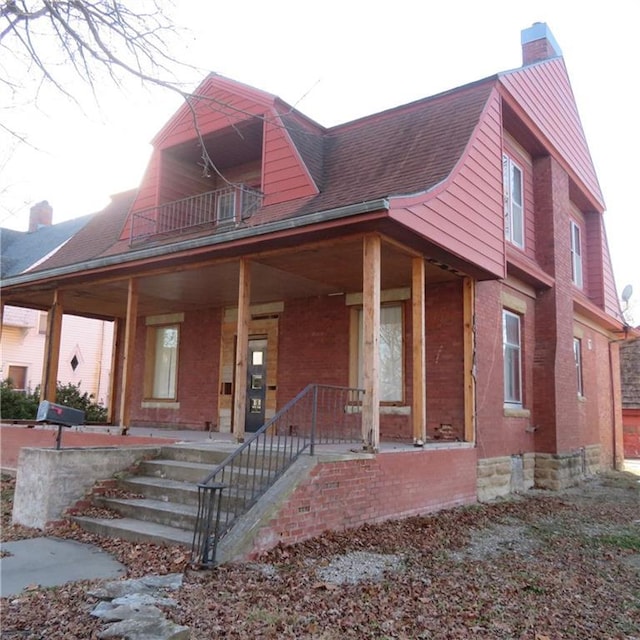 view of front of property featuring a porch, a balcony, brick siding, a shingled roof, and a chimney