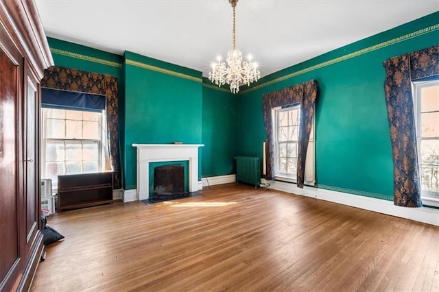 unfurnished living room featuring radiator, wood finished floors, baseboards, a fireplace with flush hearth, and a chandelier