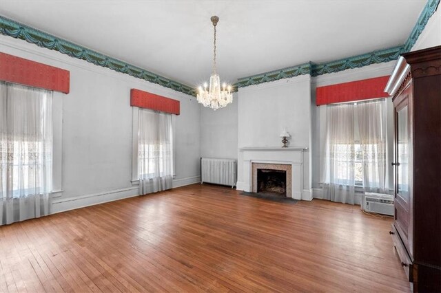 unfurnished living room with a fireplace with flush hearth, a notable chandelier, wood finished floors, radiator, and a healthy amount of sunlight