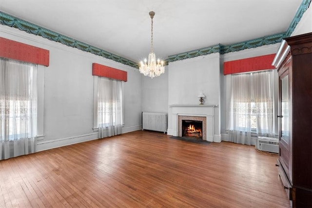 unfurnished living room with radiator, a fireplace with flush hearth, a wealth of natural light, wood finished floors, and a notable chandelier