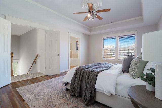 bedroom with dark hardwood / wood-style floors, connected bathroom, ceiling fan, a tray ceiling, and ornamental molding