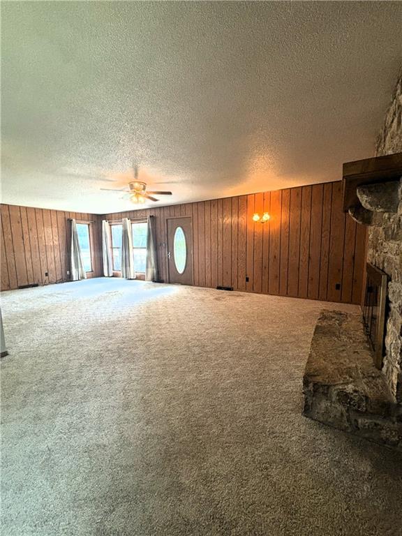 unfurnished living room with carpet, a stone fireplace, wooden walls, ceiling fan, and a textured ceiling