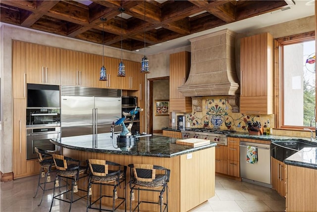 kitchen featuring stainless steel appliances, custom range hood, a sink, dark stone counters, and coffered ceiling