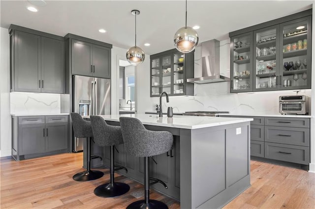 kitchen featuring an island with sink, backsplash, hanging light fixtures, wall chimney exhaust hood, and light hardwood / wood-style flooring