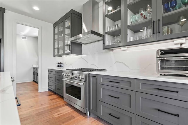 kitchen with backsplash, stainless steel range, light stone counters, light wood-type flooring, and wall chimney exhaust hood