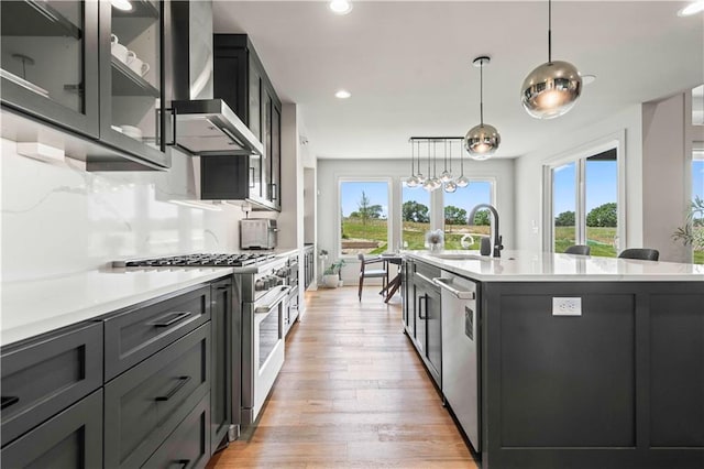 kitchen featuring appliances with stainless steel finishes, pendant lighting, sink, a center island with sink, and wall chimney range hood