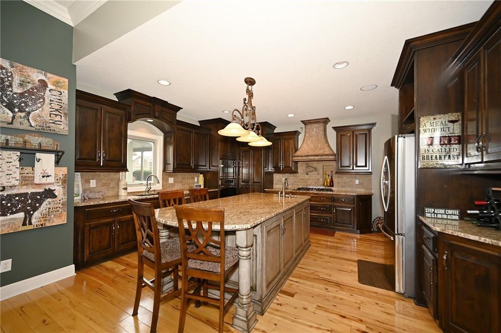 kitchen with light wood finished floors, premium range hood, a center island with sink, and decorative light fixtures