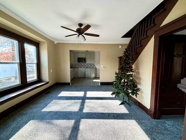 unfurnished living room with ceiling fan, crown molding, and dark colored carpet