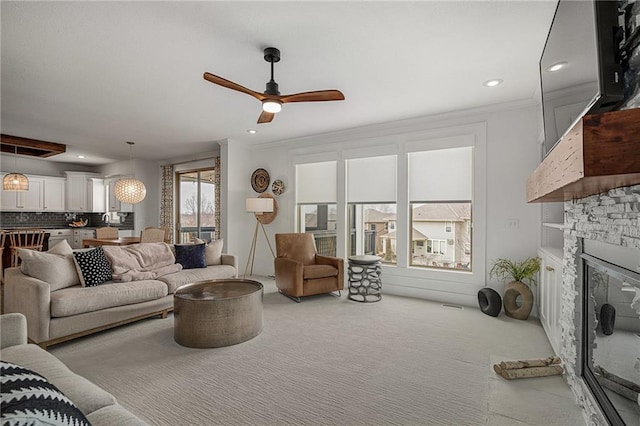 carpeted living area with a stone fireplace, recessed lighting, visible vents, a ceiling fan, and crown molding