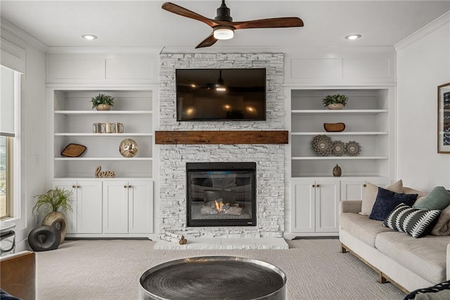 carpeted living area with ceiling fan, ornamental molding, built in features, and a stone fireplace