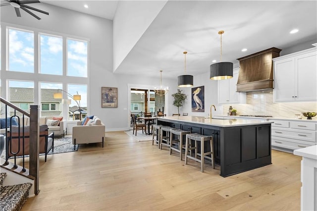 kitchen with premium range hood, ceiling fan with notable chandelier, a kitchen island with sink, decorative light fixtures, and white cabinetry