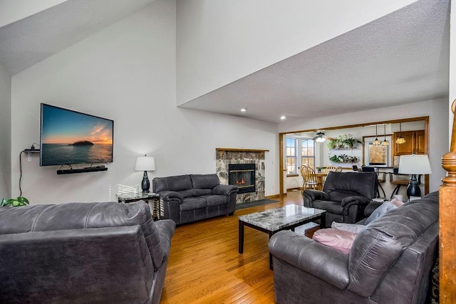 living room with a stone fireplace, vaulted ceiling, a textured ceiling, light wood-type flooring, and ceiling fan