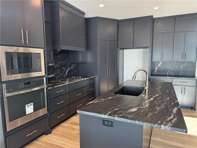 kitchen with stainless steel appliances, sink, dark stone countertops, light hardwood / wood-style floors, and a center island with sink