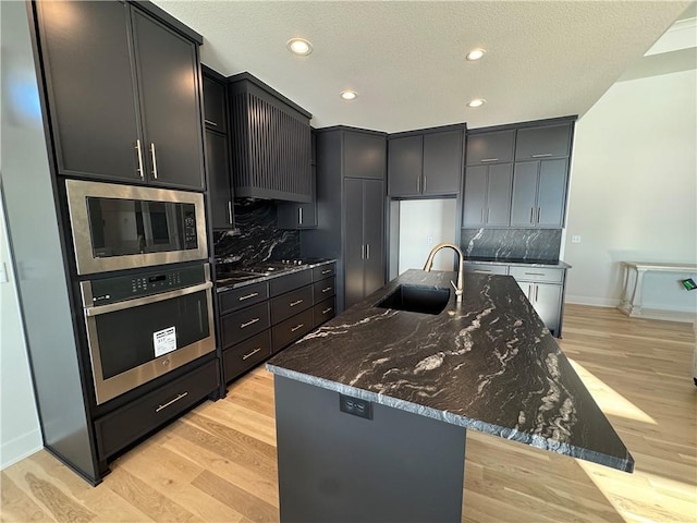 kitchen with a kitchen island with sink, light wood-type flooring, built in microwave, sink, and oven