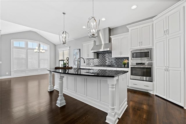 kitchen with a breakfast bar area, vaulted ceiling, an island with sink, stainless steel appliances, and wall chimney range hood